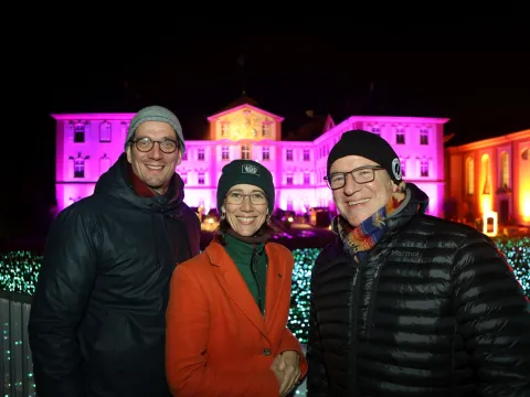 Drei Personen stehen vor einem Schloss, das bunt beleuchtet ist.