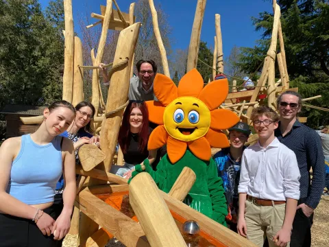 Sieben Vertreterinnen und Vertreter der Insel Mainau freuen sich zusammen mit dem Mainau-Maskottchen vor der Holz-Kugelbahn über deren Eröffnung.
