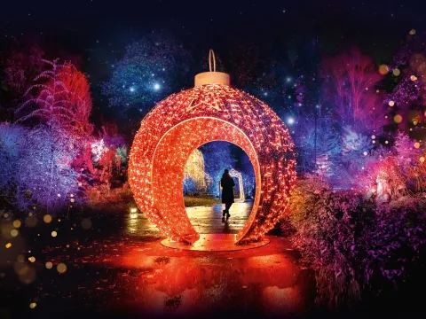 Eine rotbeleuchtete begehbare Weihnachtskugel in einer Landschaft, darin steht eine Person. Die Umgebung wird mit weihnachtlichen Lichtern angestrahlt