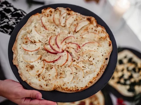Ein mit Apfel belegter Hefeteig auf einem Teller, der von zwei Händen gehalten wird. Im Hintergrund sind weitere Dinnele abgebildet
