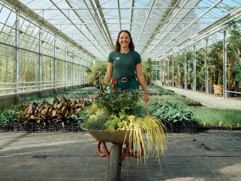 Eine Frau lacht in die Kamera. In der Hand hält sie eine Schubkarre mit grünen verschiedenen Pflanzen. Sie steht in einer Gärtnerei