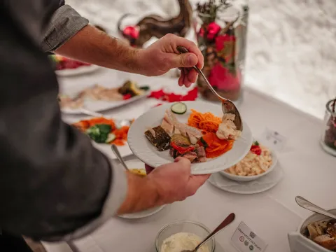 Person, die sich an einem Buffet den Teller füllt und mit einem Löffel Speisen auf einen weißen Teller gibt, auf dem bereits eingelegtes Gemüse, geraspelte Karotten, Gurkenscheiben, marinierter Fisch und Aufschnitt liegen. Der Buffettisch ist mit Schüsseln mit Salaten, Saucen und Brot gedeckt und mit roten Weihnachtsornamenten und Kerzen dekoriert, um eine festliche Atmosphäre zu schaffen.