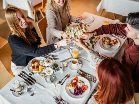 Eine Gruppe von Freunden genießt einen Brunch in einem Restaurant und erhebt ihre Champagnergläser zu einem Toast über einer weißen Tischdecke. Es werden Croissants, frisches Obst, Tomaten, Rotkohlsalat, Müsli mit Joghurt und Räucherlachs serviert, dazu Kaffee, Orangensaft und Latte Macchiato. Der Tisch ist mit einer Vase mit weißen Blumen dekoriert, was eine festliche und elegante Atmosphäre schafft