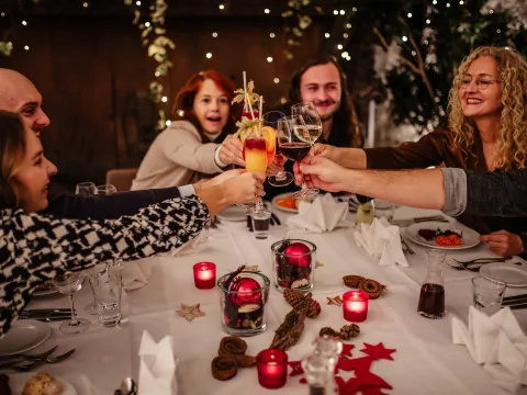 Eine Gruppe lachender Menschen sitzt an einem weihnachtlich dekorierten Tisch und stößt feierlich mit Getränken an