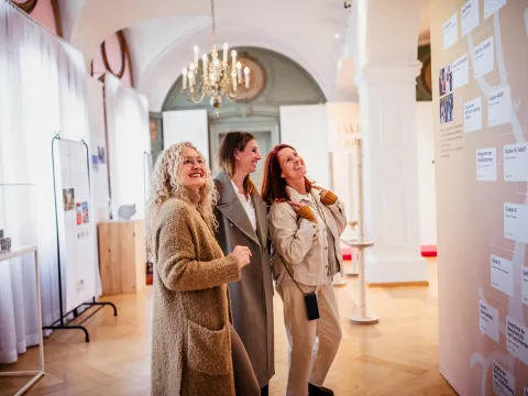 Drei lächelnde Frauen betrachten eine Ausstellung in einem hell erleuchteten Raum. Die Frauen tragen legere Mäntel und Jacken und stehen gemeinsam vor einer großen Wandtafel mit Namen und Text, wahrscheinlich einem Stammbaum oder einer historischen Zeitleiste. Der Raum ist elegant eingerichtet mit hohen Gewölbedecken, Kronleuchtern, weißen Vorhängen und Vitrinen, was eine raffinierte und kulturelle Atmosphäre schafft. Die Frauen wirken interessiert und fasziniert von der Ausstellung.  