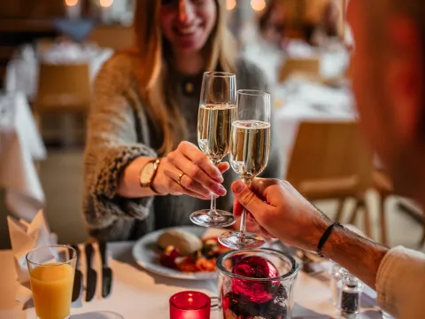 Zwei Personen stoßen mit Sektgläsern an, während sie an einem festlich gedeckten Tisch sitzen. Auf dem Tisch sind ein Teller mit Essen, ein Glas Orangensaft und eine dekorative Glasvase mit roten und schwarzen Elementen zu sehen.