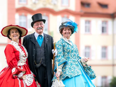 Zwei Frauen und ein Mann in historischen Kostümen lachen in die Kamera und stehen vor einem schlossähnlichem Gebäude