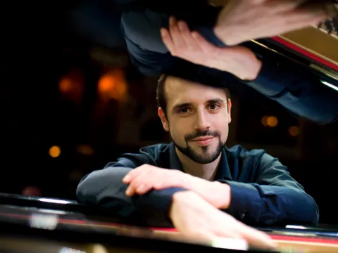 Männliche Person lehnt mit verschränkten Armen auf einem Klavier, im Hintergrund unscharfe Lichter und lächelt in die Kamera