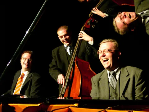 Drei Musiker in Anzügen spielen Klavier, Kontrabass und Schlagzeug in dunkler Umgebung.