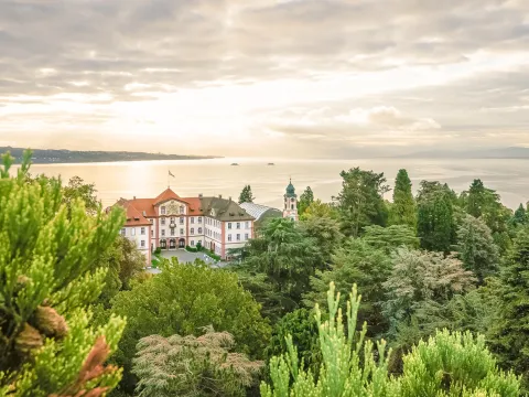 Blick über eine grüne Parklandschaft auf ein historisches Gebäude am Ufer eines Sees, im Hintergrund ruhiges Wasser und bewölkter Himmel im Abendlicht.