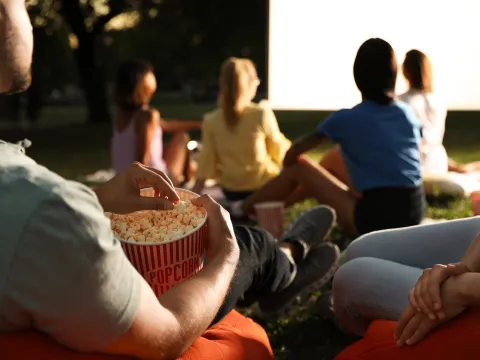 Menschen sitzen auf Sitzsäcken auf einer Wiese und schauen gemeinsam ein Open-Air-Kino, während eine Person Popcorn aus einem Becher isst.