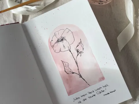 Offenes Skizzenbuch mit einer zarten Blumenzeichnung auf rosafarbenem Hintergrund, daneben Pinsel und Wasserglas
