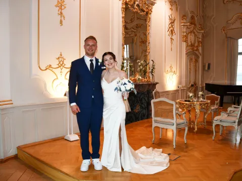 Brautpaar steht vor einem Kamin in einem prunkvollen, barock dekorierten Raum auf der Insel Mainau; die Frau trägt ein weißes Brautkleid mit Blumenstrauß, der Mann einen blauen Anzug. Im Hintergrund sind vergoldete Verzierungen, ein Spiegel, Möbel und ein Flügel zu sehen.