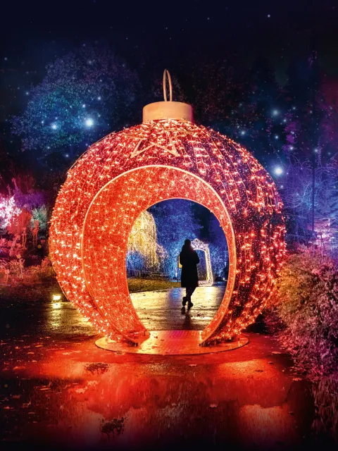 Eine rotbeleuchtete begehbare Weihnachtskugel in einer Landschaft, darin steht eine Person. Die Umgebung wird mit weihnachtlichen Lichtern angestrahlt