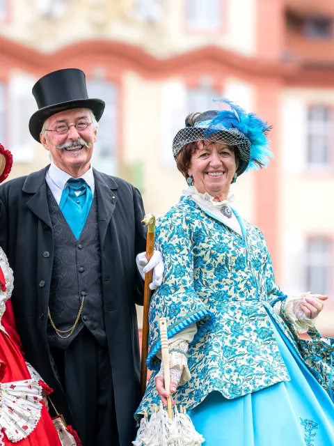 Zwei Frauen und ein Mann in historischen Kostümen lachen in die Kamera und stehen vor einem schlossähnlichem Gebäude