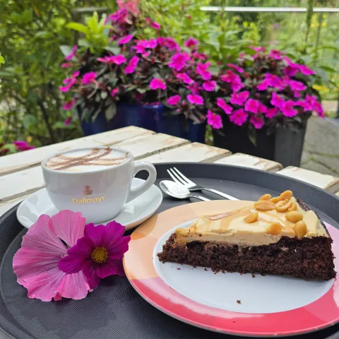 Ein Stück Schokoladenkuchen liegt auf einem Teller mit rotem Porzellanrand. Der Teller befindet sich auf einem schwarzen Tablett, ebenfalls darauf befindet sich eine Tasse Kaffee sowie eine rosa Blüte als Dekoration. Im Hintergrund sind bepflanzte Kübel und grüne Pflanzen zu sehen 